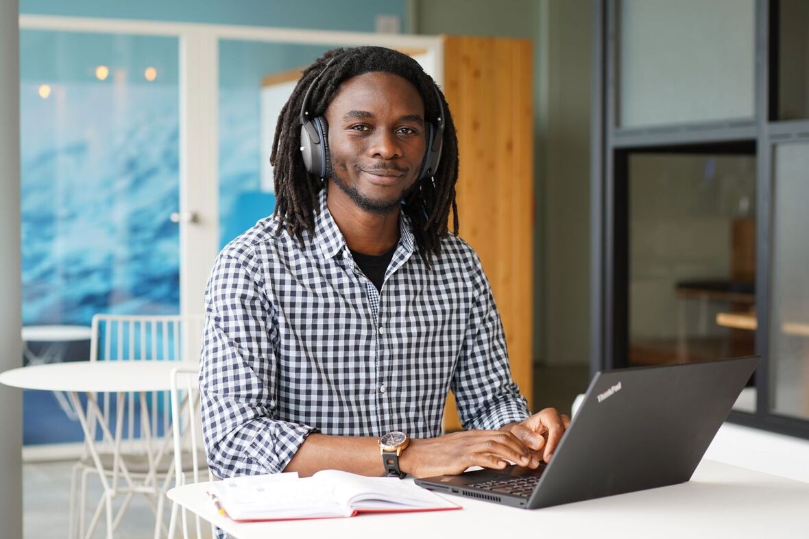 Ein junger Mann mir Bart und Dreadlocks sitzt an einem Tisch in einem Arbeitsbereich im OTTO-Bürogebäude. Neben dem Laptop liegt außerdem noch ein Notizbuch auf dem Tisch. Der Mitarbeiter hat Kopfhörer auch und schaut freundlich zur Kamera.