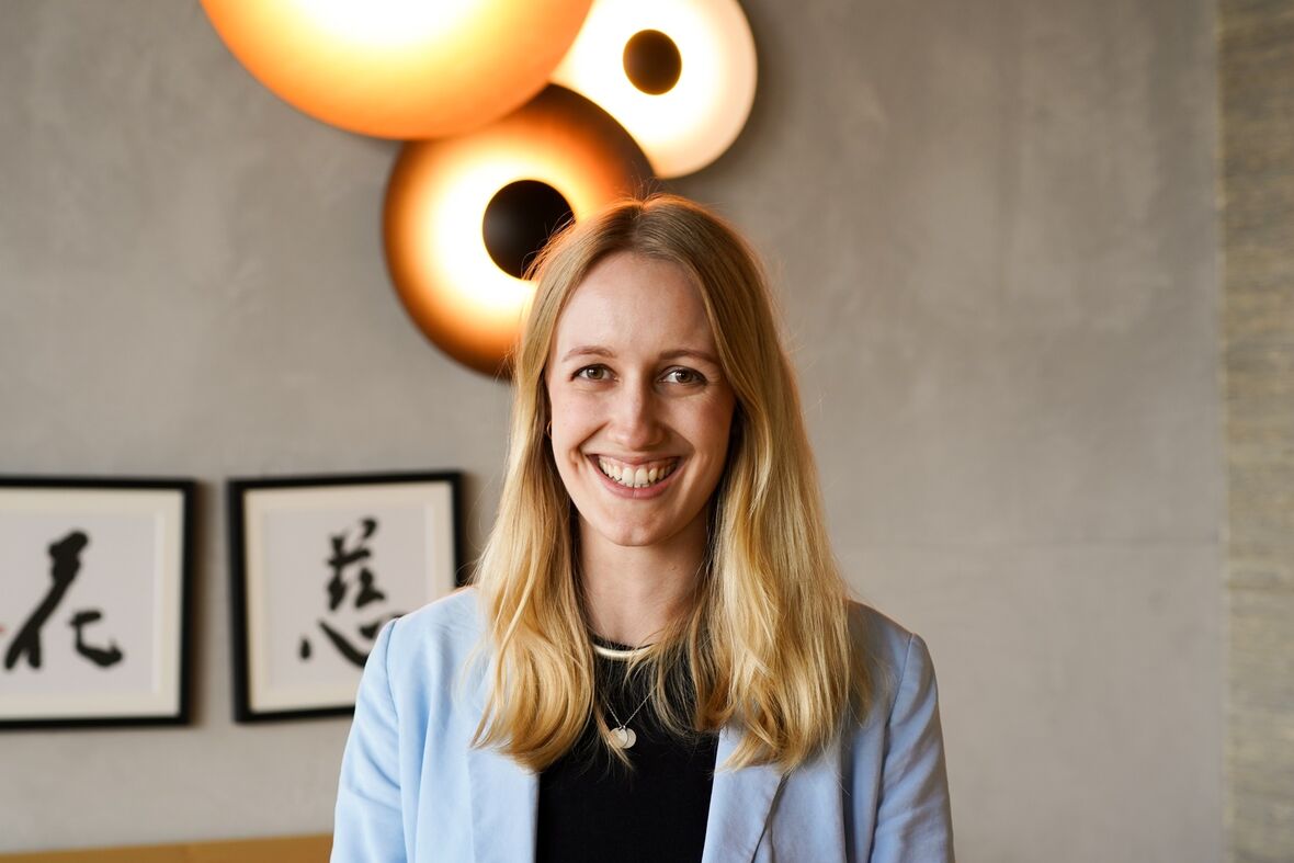 Eine Frau mit langen Haaren lächelt direkt zur Kamera. An der Wand im Hintergrund hängen moderne, kreisförmige Lampen und drei Bilder mit kalligraphierten japanischen Schriftzeichen.