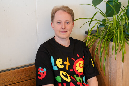 Autorenbild Nico (NiSc) Das Bild zeigt den Techblog-Autor Nico in einem grünen Pausenraum bei OTTO. Er hat blonde Haare und trägt ein buntes T-Shirt.