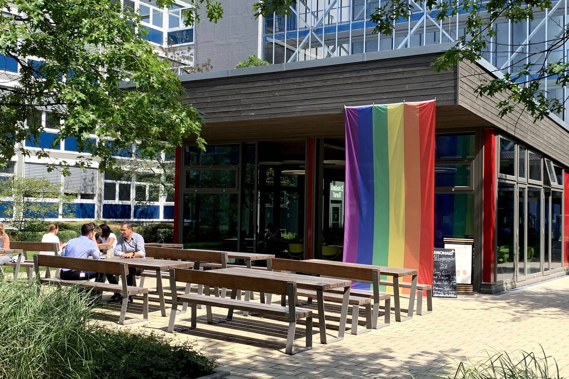 Auf dem Boulevard sind mehrere Bänke mit Tischen zu sehen, wo Mitarbeitende von OTTO sitzen. Im Hintergrund ist das kleine Gebäude des Bistros zu sehen, an dem die bunte LGTBQ-Flagge hängt. Drum herum sind mehrere Pflanzen.