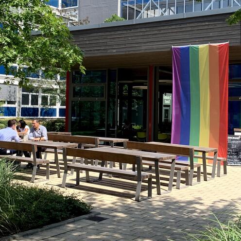 Boulevard mit Bistro Auf dem Boulevard sind mehrere Bänke mit Tischen zu sehen, wo Mitarbeitende von OTTO sitzen. Im Hintergrund ist das kleine Gebäude des Bistros zu sehen, an dem die bunte LGTBQ-Flagge hängt. Drum herum sind mehrere Pflanzen.