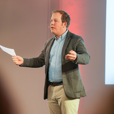 Unser Kollege hält eine Rede beim InnoBreakfast Ein Kollege steht vor einem Podium, redet in ein Mikrofon und hält in der anderen Hand einen Zettel.