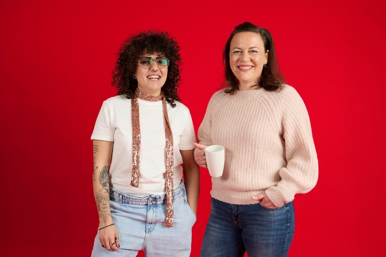 Unsere Kolleginnen stehen vor einem roten Hintergrund und lachen in die Kamera. Sie hält eine Tasse in der Hand. Die andere trägt einen rosa Schal.