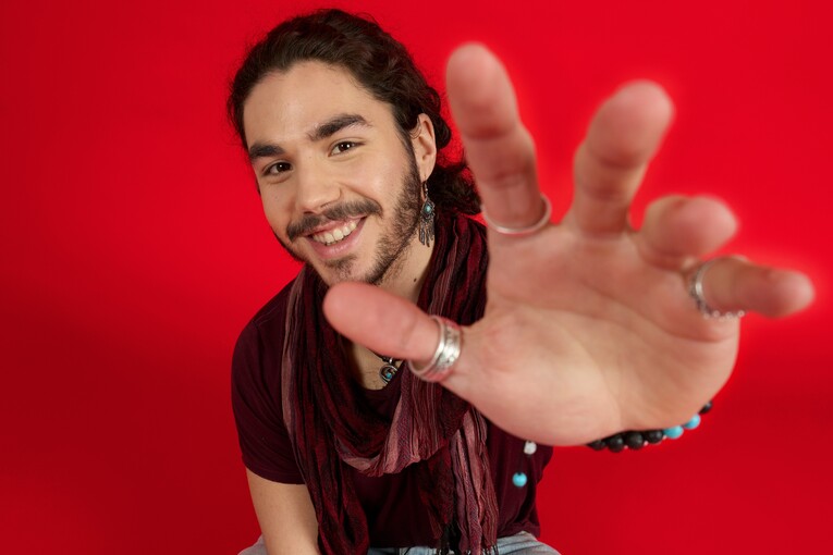 Unser Azubi in Fachinformatik sitzt im Schneidersitz auf dem Boden vor einem roten Hintergrund und lächelt in die Kamera. Mit seiner linken Hand greift er in die Kamera.