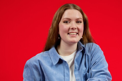 Duale Studentin in Business Administration sitzt auf einem umgedrehten Stuhl und lächelt in die Kamera. In ihrer linken Hand hält sie Kopfhörer.