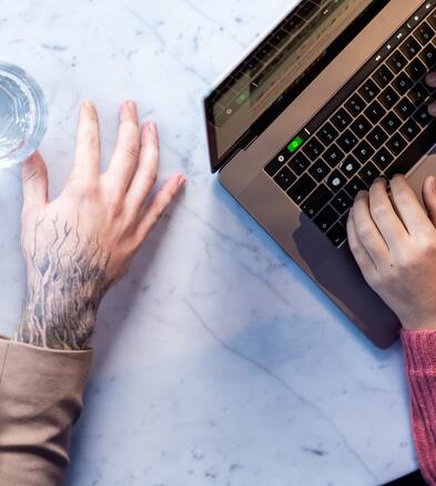 Arbeitssituation bei OTTO Zwei Kollegen sitzen nebeneinander am Tisch. Auf dem Tisch steht ein Glas Wasser und ein geöffneter Laptop. Einer von ihnen tippt auf dem Laptop. Zu sehen sind nur die Hände der Kollegen.