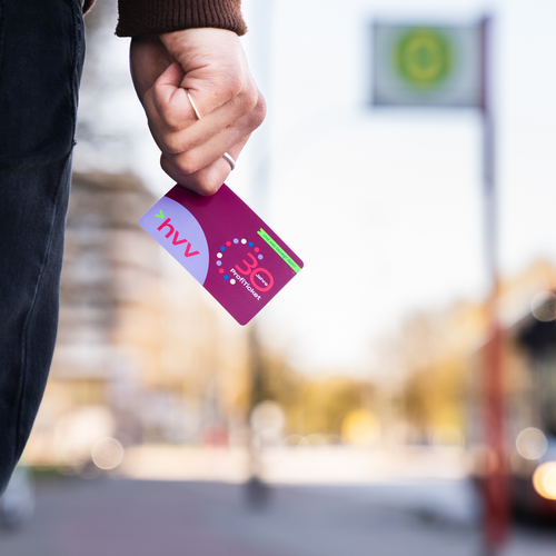 Person hält HVV-Ticket in der Hand Bild einer HVV-Karte in einer Hand vor einer verschwommenen Bushaltestelle.