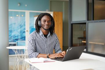 Ein junger Mann mir Bart und Dreadlocks sitzt an einem Tisch in einem Arbeitsbereich im OTTO-Bürogebäude. Neben dem Laptop liegt außerdem noch ein Notizbuch auf dem Tisch. Der Mitarbeiter hat Kopfhörer auch und schaut freundlich zur Kamera.