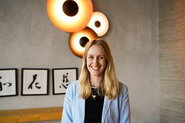 Eine Frau mit langen Haaren lächelt direkt zur Kamera. An der Wand im Hintergrund hängen moderne, kreisförmige Lampen und drei Bilder mit kalligraphierten japanischen Schriftzeichen.