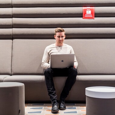 Unser Kollege Digital Analyst Ein junger Kollege, der als Digital Analyst arbeitet, sitzt mit seinem Laptop auf einem grauen Sofa in einem unserer Meetingräume und ist ganz konzentriert.