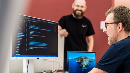 Ein IT-Mitarbeiter sitzt am Schreibtisch und guckt auf den Code, der auf dem Computerbildschirm angezeigt wird. Ein anderer Kollege steht freundlich daneben.