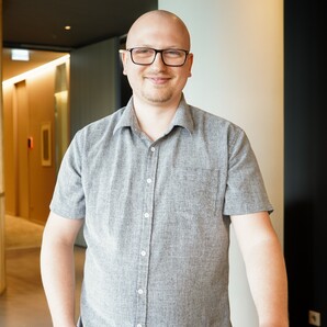 Anwendungsentwickler bei OTTO steht im Social Space. Er lächelt in die Kamera und stützt sich lässig am Tisch ab.