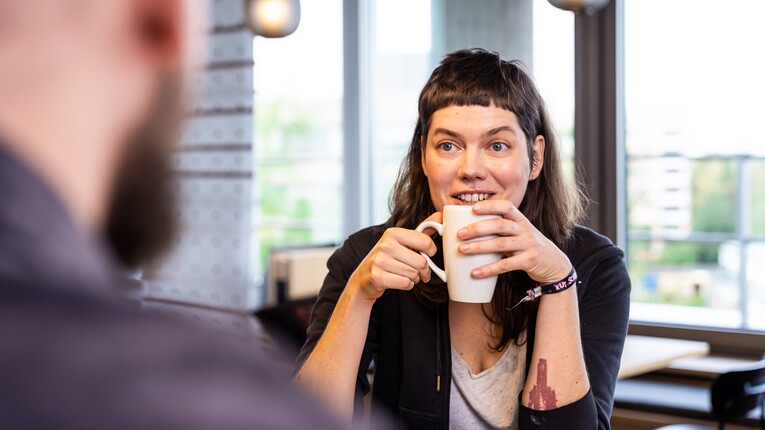 Eine Frau sitzt im Bürogebäude von OTTO einem Kollegen gegenüber. Sie hält eine Tasse in der Hand und guckt interessiert zu ihrem Gesprächspartner.