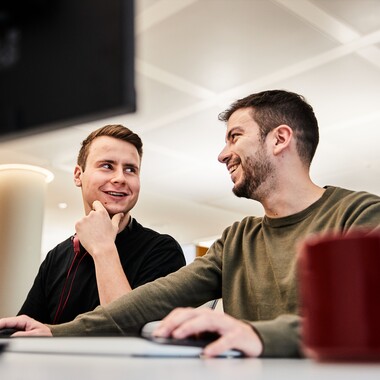 Unsere Kollegen in Tech beim Arbeiten Zwei Kollegen aus Tech sitzen gemeinsam vor dem PC und unterhalten sich lächelnd.