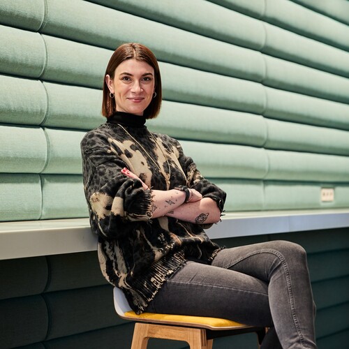 Unsere Kollegin als Venture Client Lead, Startup-Kooperationsmanagerin Unsere Kollegin sitzt mit verschränkten Armen auf dem Stuhl. und lächelt in die Kamera.