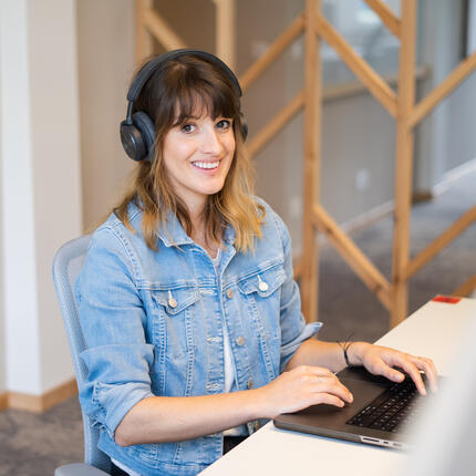 Junior Software-Entwicklerin sitzt am Schreibtisch mit Headset und tippt am Computer und lächelt in die Kamera.