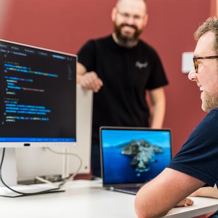 Software-Entwickler arbeiten als Team an der Weiterentwicklung unserer Plattform Ein IT-Mitarbeiter sitzt am Schreibtisch und guckt auf den Code, der auf dem Computerbildschirm angezeigt wird. Ein anderer Kollege steht freundlich daneben.