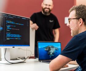Ein IT-Mitarbeiter sitzt am Schreibtisch und guckt auf den Code, der auf dem Computerbildschirm angezeigt wird. Ein anderer Kollege steht freundlich daneben.