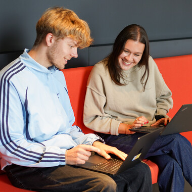 Unsere Werkstudenten sitzen am Laptop Zwei Werkstudenten eine Frau und ein Mann sitzen auf einem roten Sofa mit ihren Laptops und tauschen sich aus