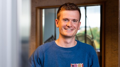 Unsere Kollege Product Manager Apps Mitarbeiter aus Projektmanagement lehnt sich an die Wand und lächelt in die Kamera.