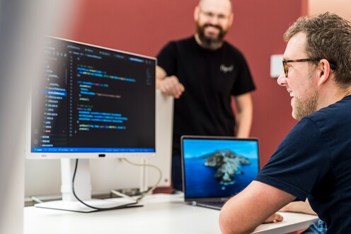 Software-Entwickler arbeiten als Team an der Weiterentwicklung unserer Plattform Ein IT-Mitarbeiter sitzt am Schreibtisch und guckt auf den Code, der auf dem Computerbildschirm angezeigt wird. Ein anderer Kollege steht freundlich daneben.