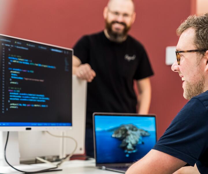 Ein IT-Mitarbeiter sitzt am Schreibtisch und guckt auf den Code, der auf dem Computerbildschirm angezeigt wird. Ein anderer Kollege steht freundlich daneben.