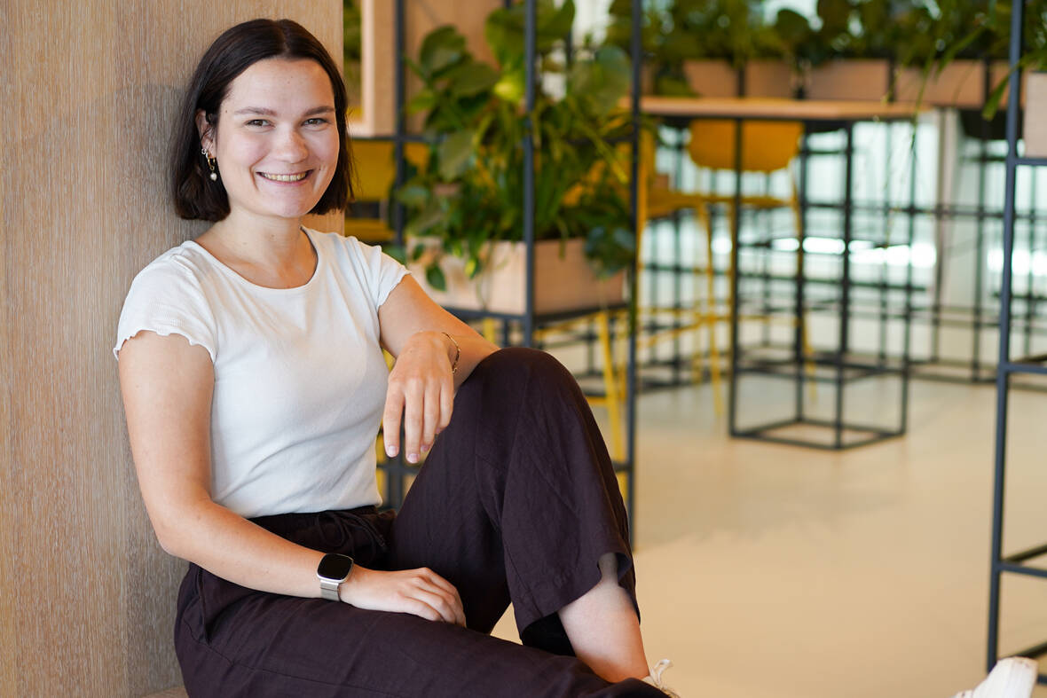 Unsere SEO Managerin sitzt angelehnt auf einer Holzbank. Ein Bein ist ausgestreckt, das andere angewinkelt. Sie lächelt in die Kamera und befindet sich in einem unserer Social-Space-Räume.
