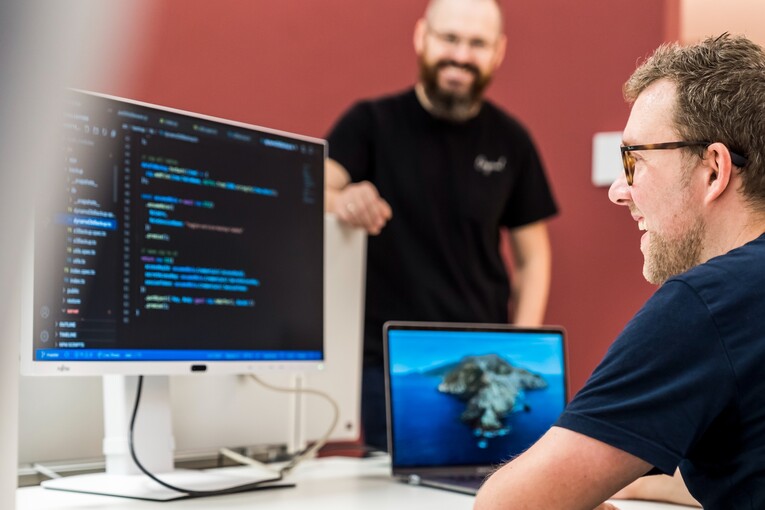 Ein IT-Mitarbeiter sitzt am Schreibtisch und guckt auf den Code, der auf dem Computerbildschirm angezeigt wird. Ein anderer Kollege steht freundlich daneben.