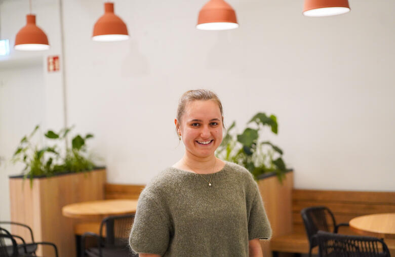 Duale Studentin in Business Administration lächelt in die Kamera Eine duale Studentin der Business Administration steht im Social Space und lächelt in die Kamera. Im Hintergrund sind mehrere Holztische und Sitzmöglichkeiten, die mit Pflanzen dekoriert sind, zu sehen.