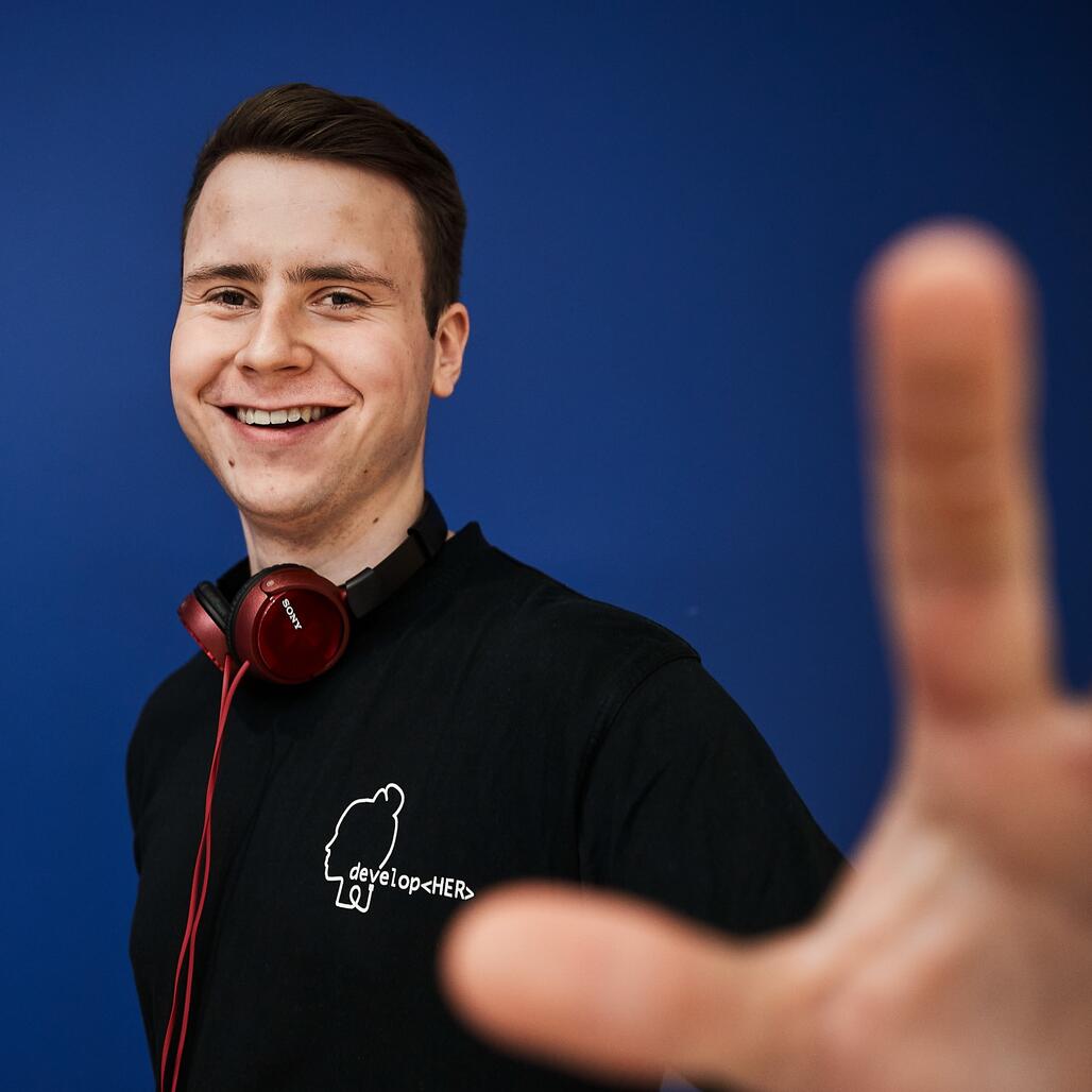 Unser Kollege steht fröhlich vor einem blauen Hintergrund und streckt den Arm nach vorne.