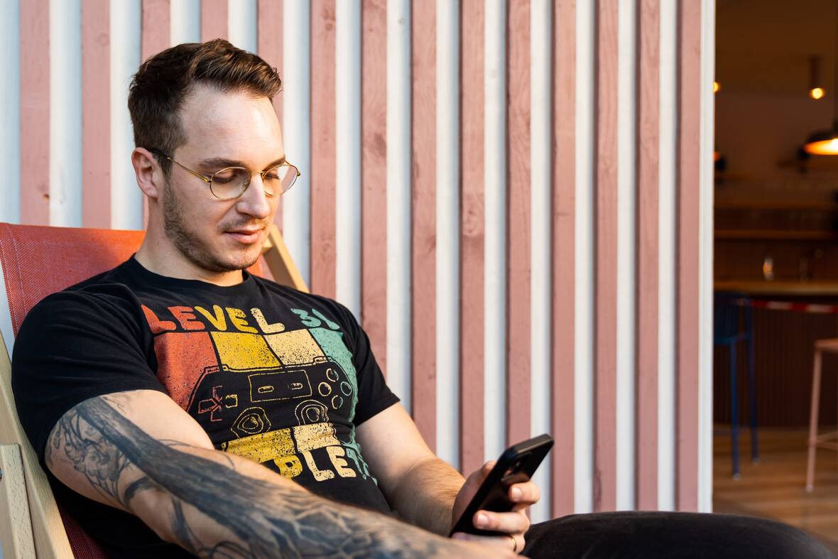 Vor einer Wand mit Holzpaneelen sitzt unser Kollege in einem Liegestuhl mit seinem Smartphone