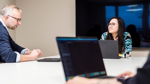 Drei OTTO-Mitarbeiter haben ein Meeting. Sie sitzen zusammen an einem Tisch im Bürogebäude und haben Laptops dabei.