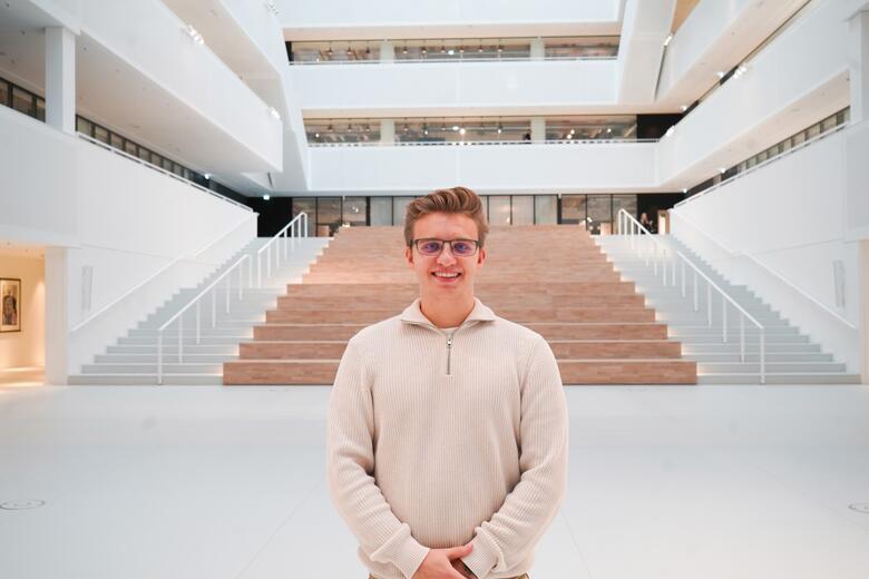 Junior Software Developer steht lächelnd im Atrium Junior Software Developer steht mittig im Atrium im neune Gebäude 4 unser Headquarter Gebäude von OTTO und lächelt in die Kamera.