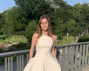 Alina, eine junge Erwachsene mit braunen Haaren, trägt ein helles Kleid und lächelt in die Kamera. Im Hintergrund ist ein Teil eines Stadtparks zu sehen. Die Sonne scheint und es sieht nach Sommer aus.