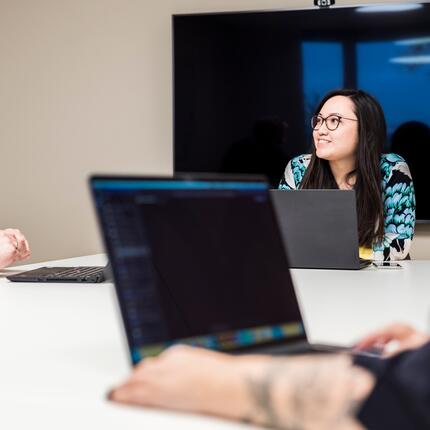 Marcel und Emmy besprechen sich in den modernen Meetingräumen Drei OTTO-Mitarbeiter haben ein Meeting. Sie sitzen zusammen an einem Tisch im Bürogebäude und haben Laptops dabei.