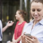 Frau guckt auf Smartphone, Kollegen stehen im Hintergrund