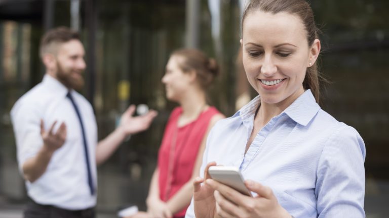 Frau guckt auf Smartphone, Kollegen stehen im Hintergrund