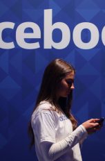 Junge Frau blickt auf Smartphone vor Wand mit Facebook-Schriftzug