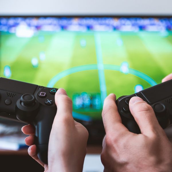 Nahaufnahme der H&auml;nde von zwei Personen, die jeweils einen Controller halten. Auf dem Monitor ist ein Fu&szlig;ballspiel zu erkennen.