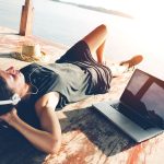 Junge Frau liegt am See und h&ouml;rt ihre Spotify-Playlist &uuml;ber ein Notebook