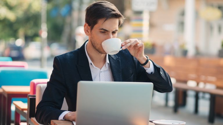 Mann sitzt kaffeetrinkend in einem Café und tippt mit angestrengtem Gesicht etwas in seinen Laptop.
