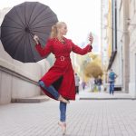 Junge Frau steht in Ballerina-Pose und mit Regenschirm auf der Stra&szlig;e und blickt auf Smartphone mit Regenradar-App