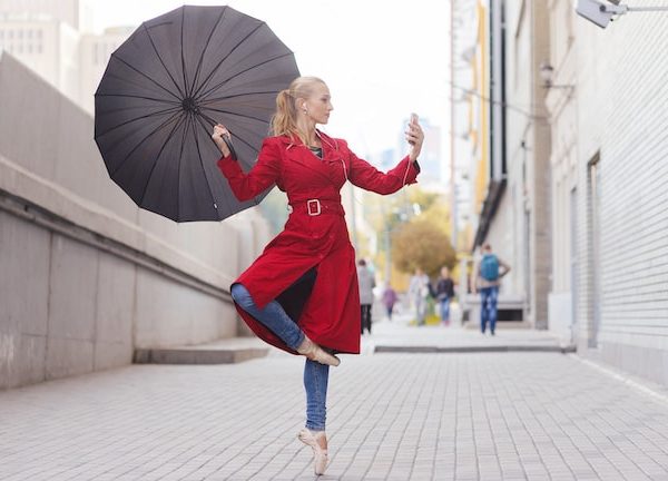 Junge Frau steht in Ballerina-Pose und mit Regenschirm auf der Straße und blickt auf Smartphone mit Regenradar-App