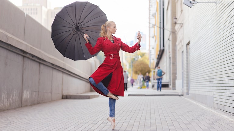 Junge Frau steht in Ballerina-Pose und mit Regenschirm auf der Straße und blickt auf Smartphone mit Regenradar-App