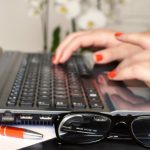 Notebook mit Hand auf Tastatur, im Vordergrund Kuli und Brille