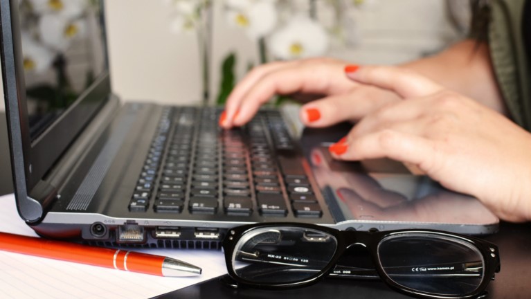 Notebook mit Hand auf Tastatur, im Vordergrund Kuli und Brille