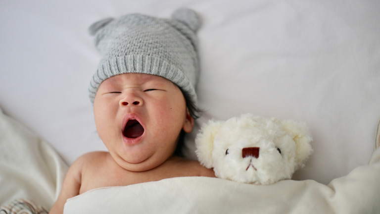 Gähnendes Baby mit Strickmütze und Teddy im Bett