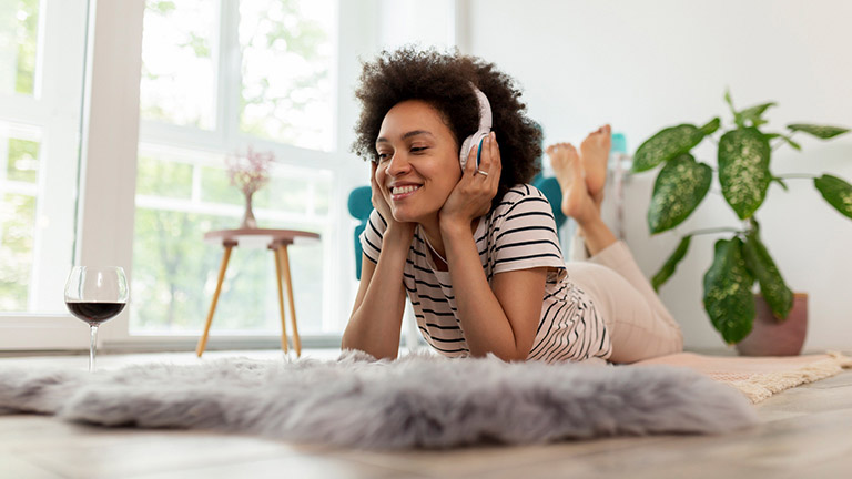 Eine Person liegt auf dem Boden und hört mit Kopfhörern Musik. In Reichweite steht ein Glas mit Rotwein.