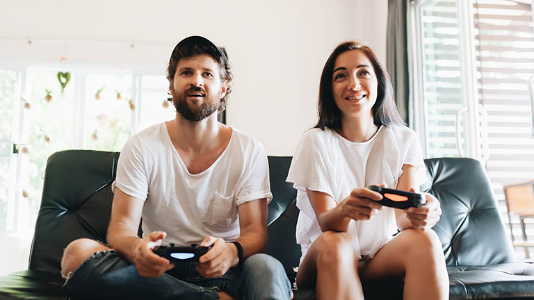 Zwei Personen sitzen auf einem Sofa und haben PS4-Controller in der Hand. Sie spielen offenbar auf einer Konsole.