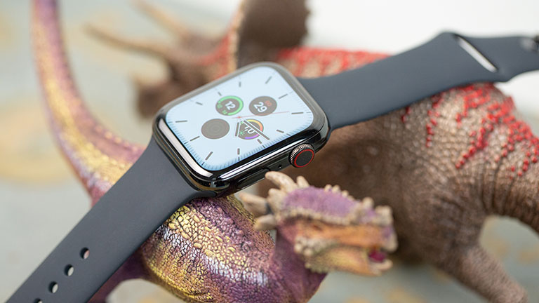 Eine Apple Watch mit schwarzem Armband ruht dekorativ auf zwei Spielzeug-Dinosaurieren. Auf dem Display ist die Uhrzeit zu lesen.